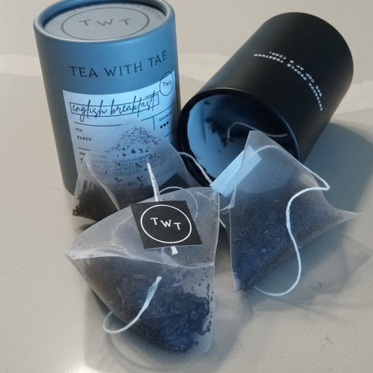 Two black cylindrical tea boxes with a clear bag containing blue tea bags on a white surface.
