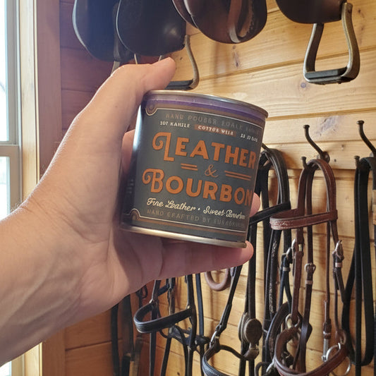 Hand holding a can of leather conditioner in front of hanging horse saddles and reins on a wooden wall.
