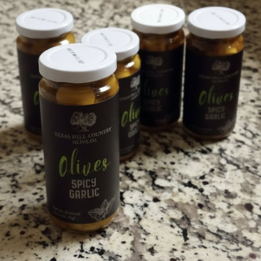 Hand holding a jar of 'Olives Spicy Garlic' with other similar jars on a marble countertop.