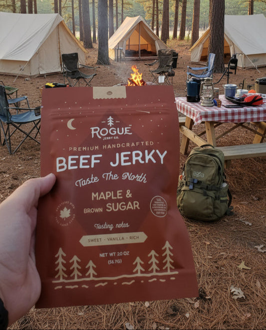 Person holding a package of Rogue Beef Jerky in a forest setting with tents and campfire.