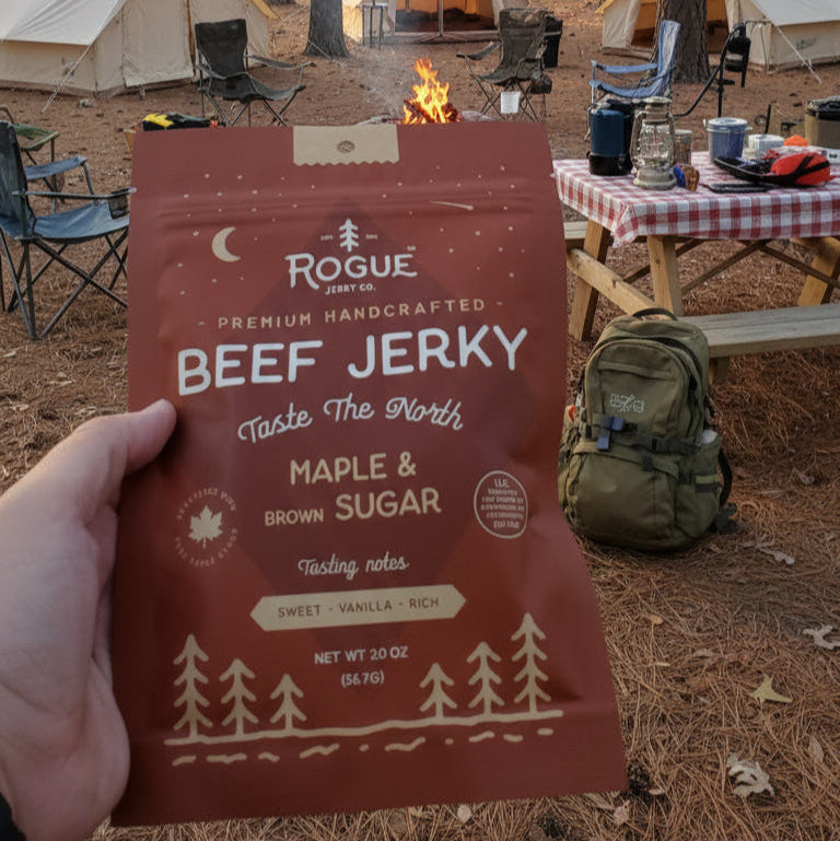 Person holding a package of Rogue Beef Jerky in a forest setting with tents and campfire.