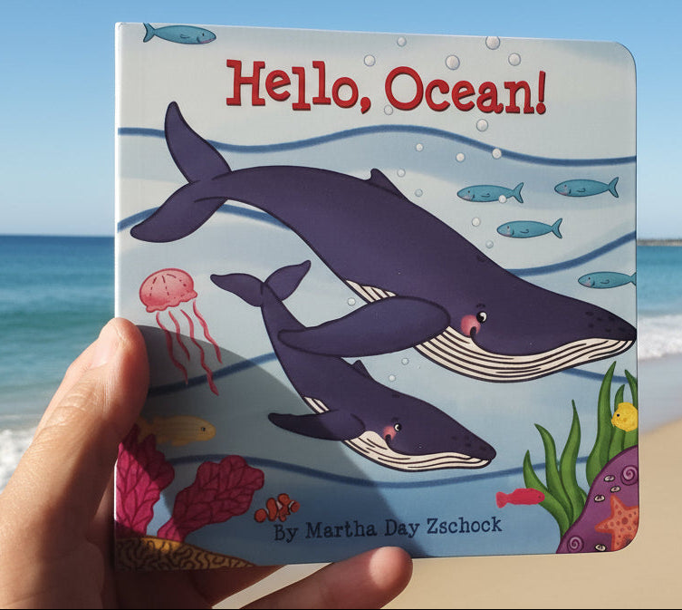 Children's book titled 'Hello, Ocean!' held by a hand on a wooden surface