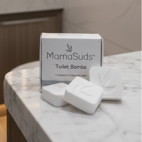 Three white MamaSuds toilet bomb bars arranged in front of their white and silver packaging box on a white ceramic surface.