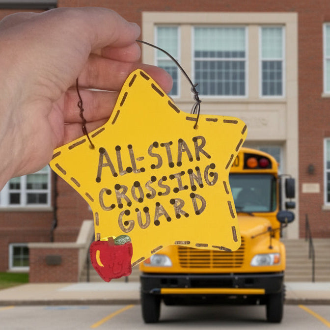 Hand holding a 'All-Star Crossing Guard' sign in front of a school bus with a school building in the background.