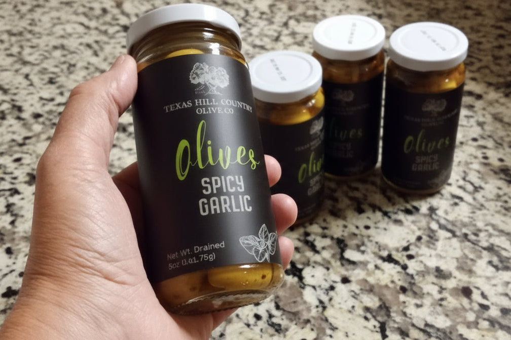 Hand holding a jar of 'Olives Spicy Garlic' with other similar jars on a marble countertop.