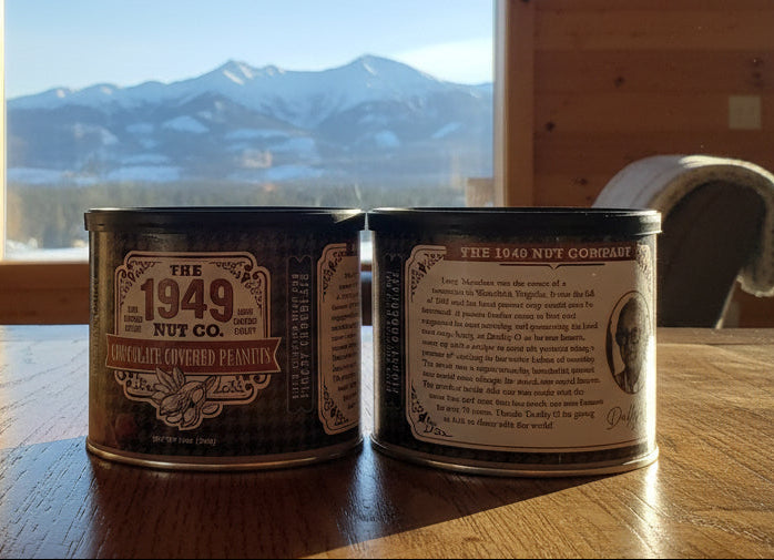 Two cans of 'The 1949 Nut Co.' on a wooden table with a mountain view in the background.