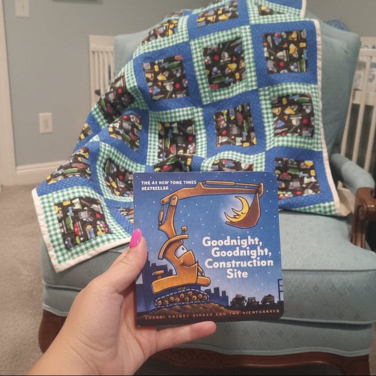 Child's quilt with construction site pattern held by a hand in a room with a crib and baby monitor.